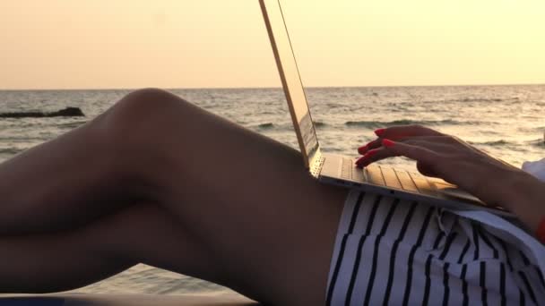 Close-up, pernas femininas em shorts. jovem usa laptop, deitado em uma espreguiçadeira, na praia junto ao mar, ao pôr do sol ou ao nascer do sol. freelancer, trabalho remoto. férias de verão no mar — Vídeo de Stock