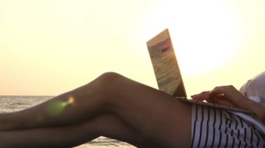Yakın plan, şortlu kadın bacakları. Genç kadın dizüstü bilgisayar kullanıyor, deniz kıyısında, gün batımında ya da gün doğumunda bir şezlongda yatıyor. Serbest çalışan, uzaktan kumandalı. Denizde yaz tatili