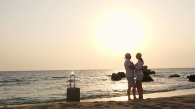 Balayı. Romantik aşıklar, siluetler. Gezginler sahilde durur, denizin arka planına sarılır ve öpüşürler, güneşin doğuşuna veya batışına karşı. Yakınlarda bavul var. seyahat konsepti. deniz kenarı tatili.