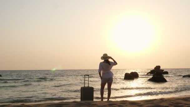 サンセットや日の出に海のビーチでスーツケースを着たリラックスした女性のシルエット。旅行のコンセプト. — ストック動画