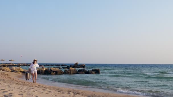 Młoda kobieta w krótkich spodenkach, kapeluszu i okularach przeciwsłonecznych, nosi walizkę w jednej ręce, torebkę i sandały w drugiej. powoli, zmęczenie spaceruje wzdłuż plaży nad morzem. koncepcja nieprzewidzianych przygód na morzu — Wideo stockowe