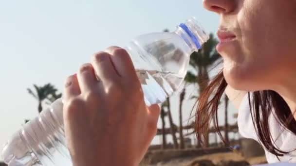 Gros plan, femme en lunettes de soleil, chapeau de soleil, boit l'eau d'une bouteille en plastique sur fond de palmiers, par une chaude journée d'été. — Video