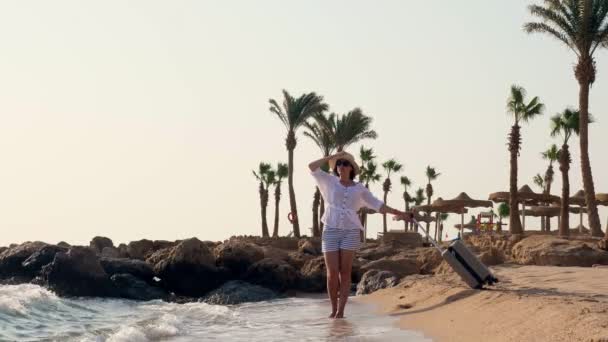 Mujer joven en ropa de verano y sombrero de sol, sosteniendo bolsa de viaje, mirando al mar, disfrutando del clima, sobre el fondo de la playa y las palmeras. — Vídeos de Stock