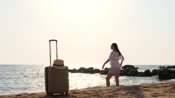 日の出に腕を伸ばして海のそばのビーチで自由と人生を楽しむ若い女性。近くに旅行用のスーツケースがある。旅行と健康の概念 — ストック動画