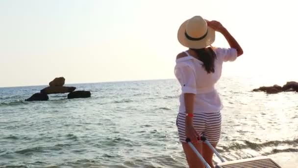Widok z tyłu młodej kobiety w letnich ubraniach i kapeluszu, trzymając torbę podróżną, patrząc na morze, ciesząc się wschodem słońca, koncepcja podróży — Wideo stockowe