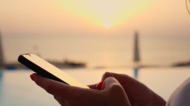 Yakın plan, cep telefonu ve kadın eli, havuzun arka planına ve gündoğumuna karşı. Kadınlar akıllı telefon kullanır.