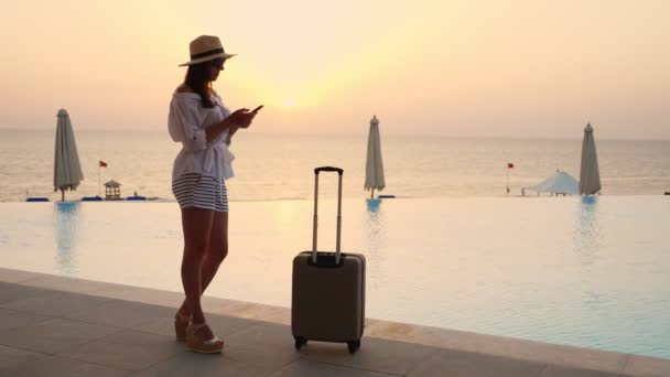 Νεαρή γυναίκα με καλοκαιρινά ρούχα και καπέλο, με ταξιδιωτικές αποσκευές, χρησιμοποιώντας smartphone, ενώ στέκεται στην πισίνα, την ανατολή του ηλίου. Καλοκαίρι. παραθαλάσσιο θέρετρο. ταξιδιωτική έννοια. — Αρχείο Βίντεο