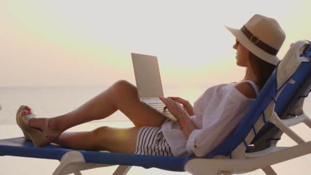 戴着太阳镜和太阳帽的年轻女子，在日落或日出时躺在躺椅上使用笔记本电脑。自由职业者，远程工作。在海滨度假胜地的暑假. — 图库视频影像