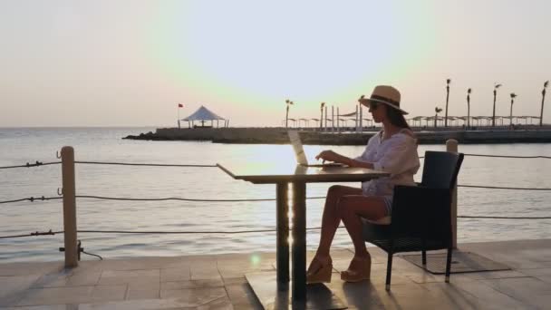 Junge Frau in Sommerkleidung, Sonnenbrille und Sonnenhut, benutzt Laptop, sitzt allein am Tisch an der Strandpromenade, bei Sonnenuntergang oder Sonnenaufgang. Freiberufler, Fernarbeit. Sommerurlaub auf See — Stockvideo