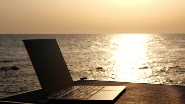 Close-up, ένα ανοικτό φορητό υπολογιστή βρίσκεται στο τραπέζι στο φως του ήλιου, με φόντο την ανατολή του ηλίου ή το ηλιοβασίλεμα από τη θάλασσα. — Αρχείο Βίντεο