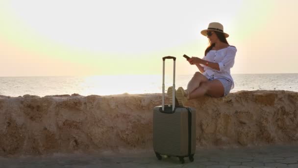 夏の服、サングラス、太陽の帽子の若い女性は、旅行用の荷物を持って、空の遊歩道で、海のそばに立っている。海辺での休暇。旅のコンセプト — ストック動画
