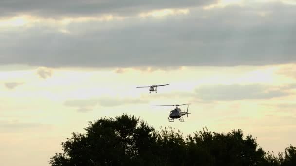 Avião retro e um helicóptero estão voando sobre as copas das árvores, contra o céu por do sol com nuvens escuras de tempestade. raios solares estão rompendo através das nuvens — Vídeo de Stock