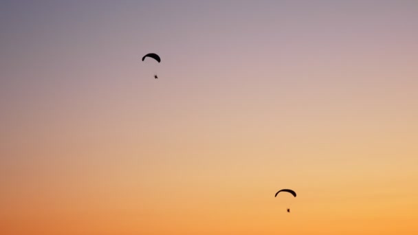 Siluetas oscuras de dos Moto Parapentes vuelan, se elevan en el aire, sobre el fondo de una puesta de sol naranja brillante — Vídeo de stock