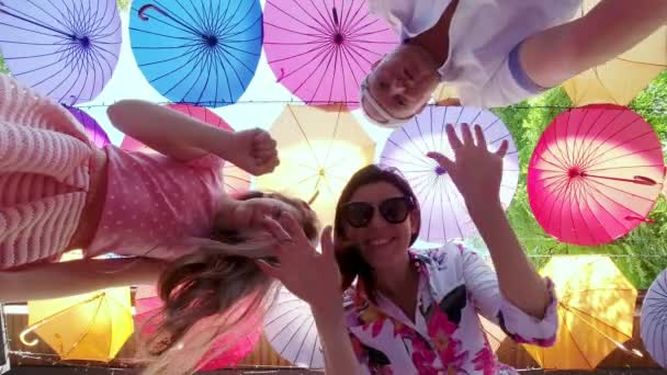 Famille de trois, maman, papa et fille, prendre selfie, sur fond de parasols décoratifs multicolores suspendus. ils dansent et s'amusent tout en filmant la vidéo. — Video
