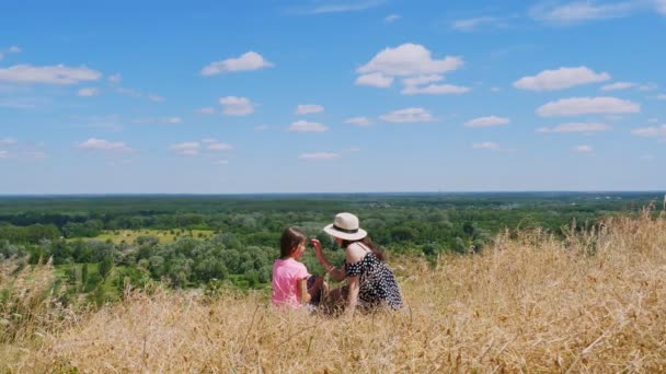 Beautiful summer landscape. woman and little girl, mom and daughter, sit on cliff edge, talking and admiring amazing panorama of green valley, and blue sky with rare clouds. Back view. — Stock Video