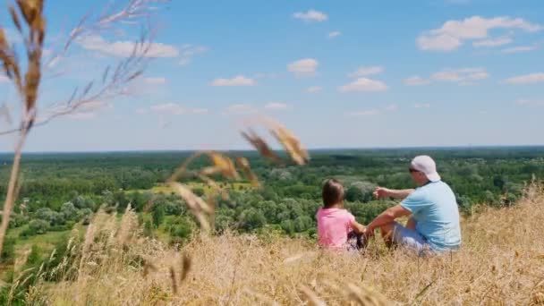 Krásná letní krajina. muž a malá holčička, otec a dcera, sedí na okraji útesu, mluví a obdivují úžasné panorama zeleného údolí a modrou oblohu se vzácnými mraky. Zadní pohled. — Stock video