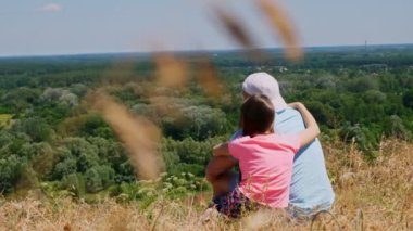 Güzel yaz manzarası. Bir adam ve küçük bir kız, baba ve kız uçurumun kenarında oturuyor, konuşuyor ve hayranlık verici yeşil vadi manzarası, ve ender bulutlarla mavi gökyüzü. Arka plan.