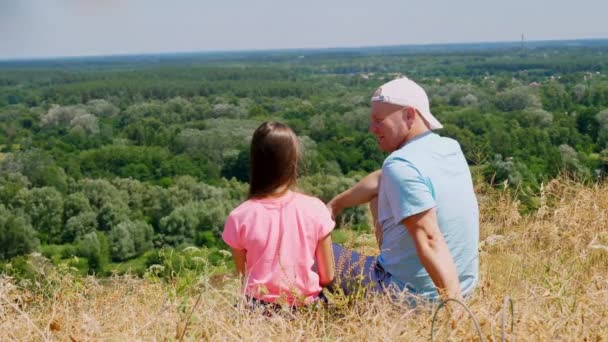Krásná letní krajina. muž a malá holčička, otec a dcera, sedí na okraji útesu, mluví a obdivují úžasné panorama zeleného údolí a modrou oblohu se vzácnými mraky. Zadní pohled. — Stock video