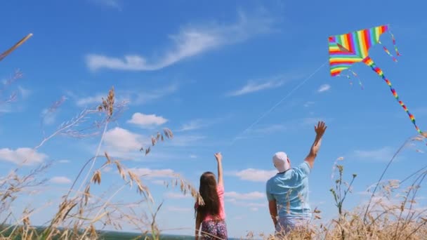 Mutlu aile, baba ve kız, mavi gökyüzünün arka planında dikenli bir alanda duran çok renkli bir uçurtma uçuruyorlar. Güneşli yaz günü. — Stok video