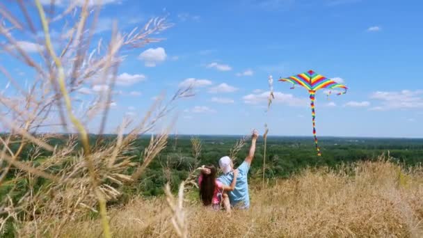 Gelukkige familie, vader en dochter, zitten op klif rand, vliegen een multi-gekleurde vlieger, plezier hebben, tegen de achtergrond van een prachtig panorama van groene vallei, en blauwe lucht met zeldzame wolken. Achteraanzicht. zonnige dag — Stockvideo