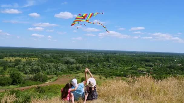 Šťastná mladá rodina sedí na okraji útesu, létat pestrobarevný drak, baví, na pozadí úžasné zelené údolí panorama, a modré nebe se vzácnými mraky. Zadní pohled. slunečný den — Stock video