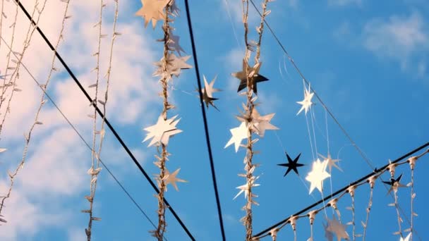 Close-up, garlands of light bulbs and silver stars, sway in the wind, glitter in the suns rays. Light street decoration against the blue sky. — Stock Video