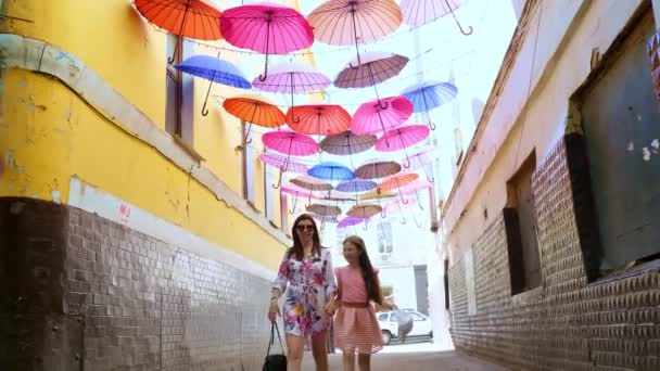 Egy fiatal nő egy tinédzser lánnyal sétálgat az utcán színes esernyőkkel díszítve. utcai dekoráció sokszínű esernyőkkel — Stock videók