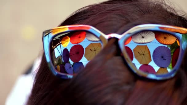 Yakın plan, siyah güneş gözlükleriyle, sokak dekorasyonundan gelen çok renkli şemsiyeler yansıtılıyor — Stok video