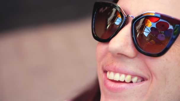 Nahaufnahme, schönes, lächelndes weibliches Gesicht in Sonnenbrille, in der sich bunte Regenschirme aus der Straßendekoration spiegeln — Stockvideo
