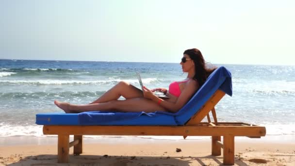 Pembe mayo ve güneş gözlüğü takan genç bir kadın dizüstü bilgisayar kullanıyor, deniz kenarında, sıcak yaz gününde uzanmış yatıyor. Serbest çalışan, uzaktan kumandalı. Denizde yaz tatili — Stok video