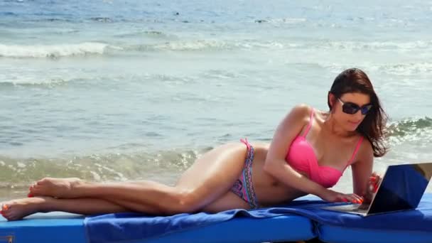 Mujer joven en traje de baño de color rosa brillante y gafas de sol, utiliza el ordenador portátil, acostado en la tumbona, en la playa junto al mar, día caluroso de verano. freelancer, trabajo remoto. vacaciones de verano en el mar — Vídeos de Stock