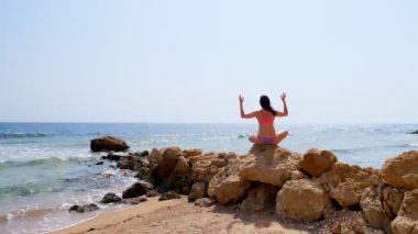 Arkadan bak. Mayolu genç bir kadın deniz kıyısındaki taşın üzerinde otururken yoga yapıyor. vücut bakımı ve meditasyon kavramı