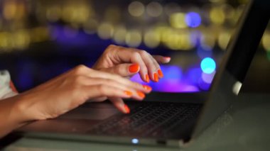 Yakın plan, parlak kırmızı tırnaklı kadın elleri, bir dizüstü bilgisayarın klavyesinde, bulanık şehir gece lambaları arka planında.