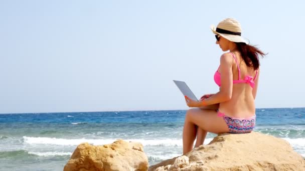 Jovem mulher em maiô, chapéu de sol e óculos de sol, usa laptop, sentado em pedra na praia junto ao mar, dia quente de verão. freelancer, trabalho remoto. férias de verão no mar — Vídeo de Stock