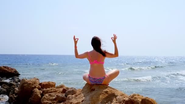 Vista para trás. jovem mulher em maiô medita, praticando ioga, enquanto sentado em pedra na praia junto ao mar. cuidado do corpo e conceito de meditação — Vídeo de Stock