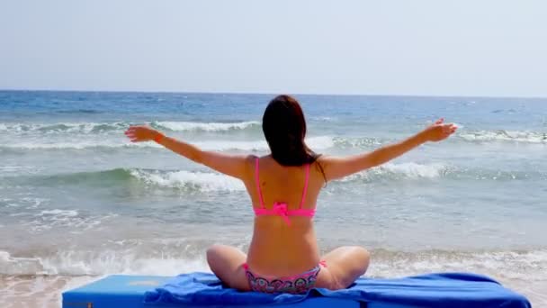 Vista trasera. mujer joven en traje de baño medita, practicando yoga, mientras está sentada en una tumbona, en la playa junto al mar. concepto de cuidado corporal y meditación — Vídeos de Stock