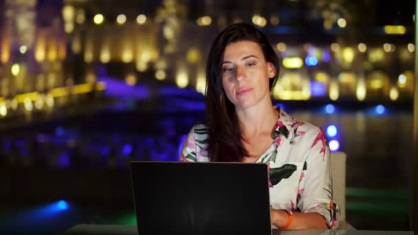 Piękna młoda brunetka używa laptopa, siedzi przy stole na otwartym tarasie lub balkonie, na tle nocnego miasta jasne światła. ciepła letnia noc. — Wideo stockowe