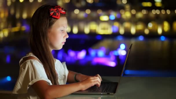 Carino teen girl utilizza il computer portatile, seduto a tavola sulla terrazza aperta o balcone, sullo sfondo della città di notte luci luminose. calda notte d'estate. — Video Stock