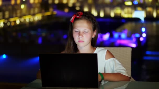 Nettes Teenie-Mädchen benutzt Laptop, sitzt am Tisch auf offener Terrasse oder Balkon, vor dem Hintergrund der hellen Nachtbeleuchtung der Stadt. Warme Sommernacht. — Stockvideo