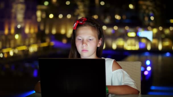 Bonito adolescente menina usa laptop, sentado à mesa no terraço aberto ou varanda, contra o fundo da noite da cidade luzes brilhantes. noite quente de verão . — Vídeo de Stock