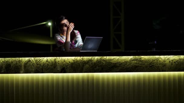 Красивая молодая брюнетка использует ноутбук, сидя за столом на открытой террасе или балконе, на фоне ночного города яркие огни. тёплая летняя ночь. — стоковое видео