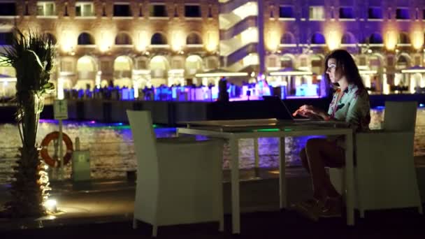 Bela jovem morena usa laptop, sentado à mesa no terraço aberto ou varanda, contra o fundo da cidade noturna luzes brilhantes. noite quente de verão . — Vídeo de Stock