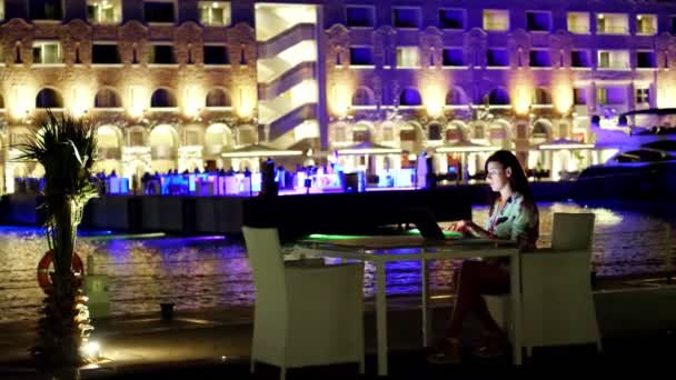 Piękna młoda brunetka używa laptopa, siedzi przy stole na otwartym tarasie lub balkonie, na tle nocnego miasta jasne światła. ciepła letnia noc. — Wideo stockowe