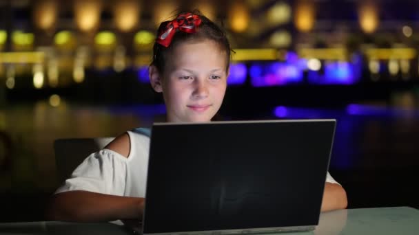 Linda chica adolescente utiliza el ordenador portátil, sentado en la mesa en la terraza o balcón abierto, contra el fondo de las luces brillantes de la noche de la ciudad. noche cálida de verano. — Vídeo de stock
