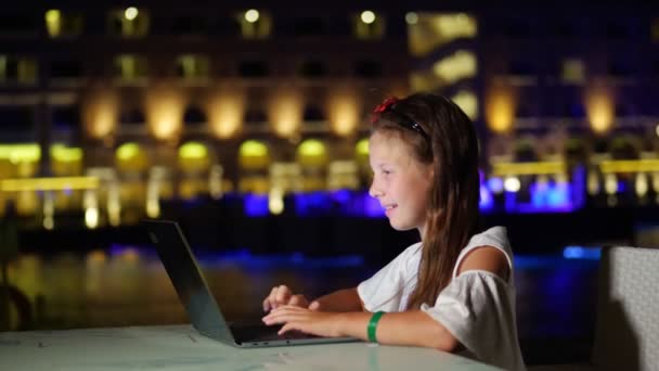 Söt tonåring flicka använder laptop, sitter vid bordet på öppen terrass eller balkong, mot bakgrund av natten staden ljus. varm sommarnatt. — Stockvideo