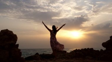 Arka manzara, gün doğumunda denizin kenarında duran genç bir kadının karanlık silueti. Rüzgar elbisesini üfledi.