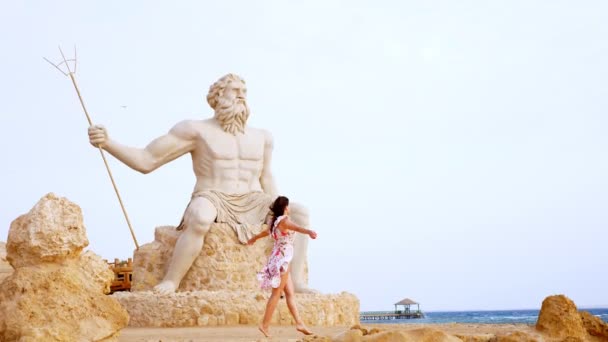 Seorang wanita muda yang cantik dalam gaun ringan, berkibar dalam angin, berjalan di sepanjang pantai berbatu. terhadap latar belakang patung monumental Allah Poseidon. — Stok Video