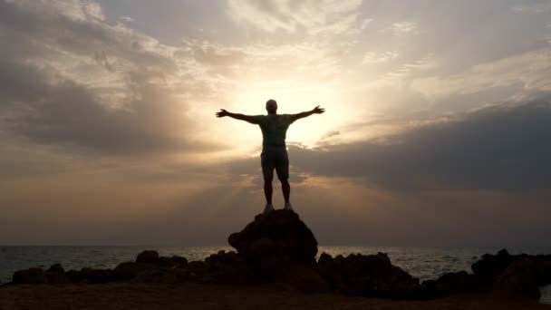 Dark male silhouette, man stands on rocks in front of the ocean or sea, with his arms outstretched and hands up at dawn, sunrise. concept of human strength and faith — Stock Video
