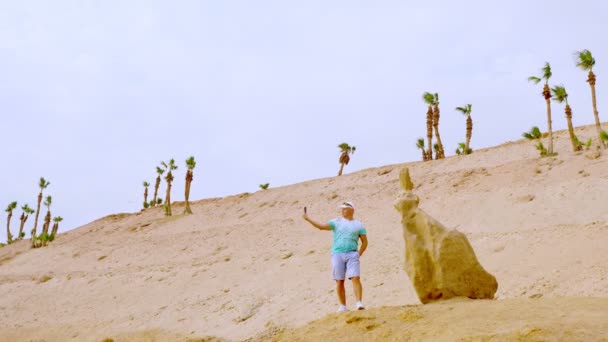 携帯電話で話してるハンサムな陸上競技の男セルフィーを取ってオンラインビデオ放送をリードしてる。砂漠を背景に大きな石のそばに一本のヤシの木があります。ブロガーはビデオを録画しています , — ストック動画