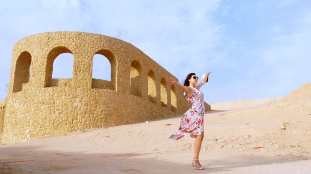 Sonriente, hermosa joven, con gafas de sol y vestido de verano ligero, revoloteando en el viento, se encuentra entre el desierto, sobre el fondo de un edificio arquitectónico, en un día caluroso de verano — Vídeo de stock